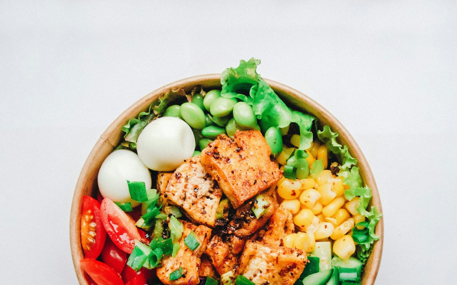 A bowl of grilled tofu, boiled eggs, sweetcorn and greens on a white table